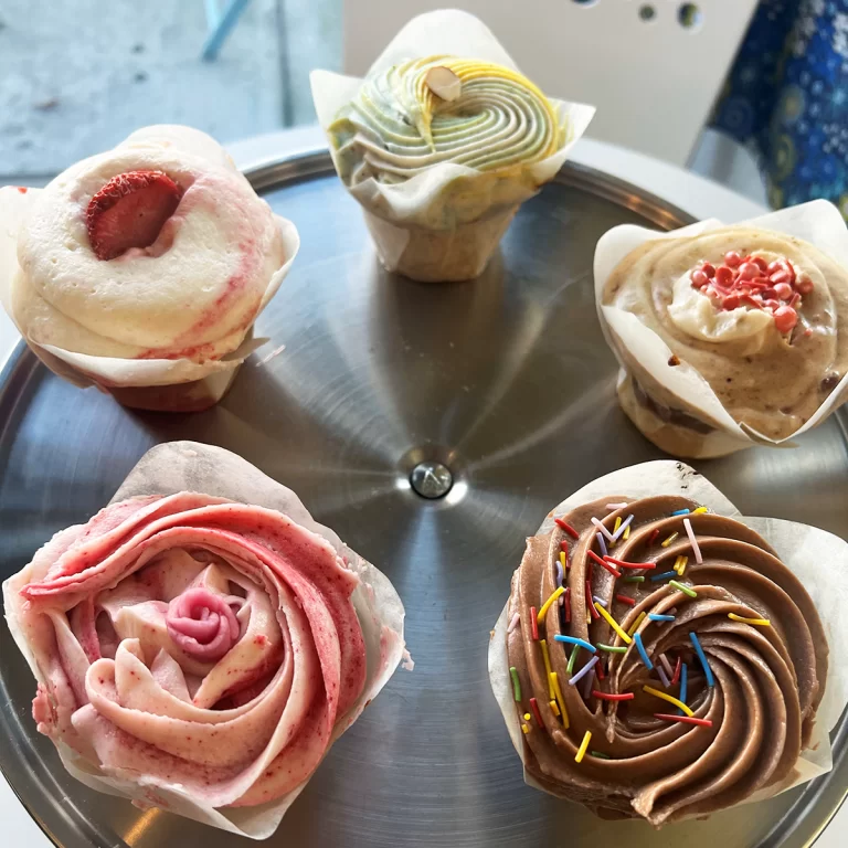 Five assorted cupcakes with different frosting designs and colors on a metal tray.